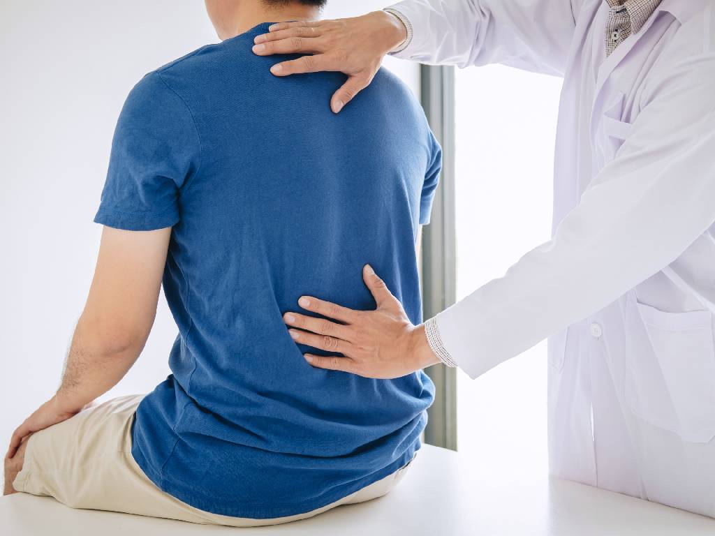 A view from behind of a patient in a blue t-shirt sitting on an examination table while a medical professional in a white lab coat performs a physical assessment, placing one hand on the upper back and the other on the lower back.