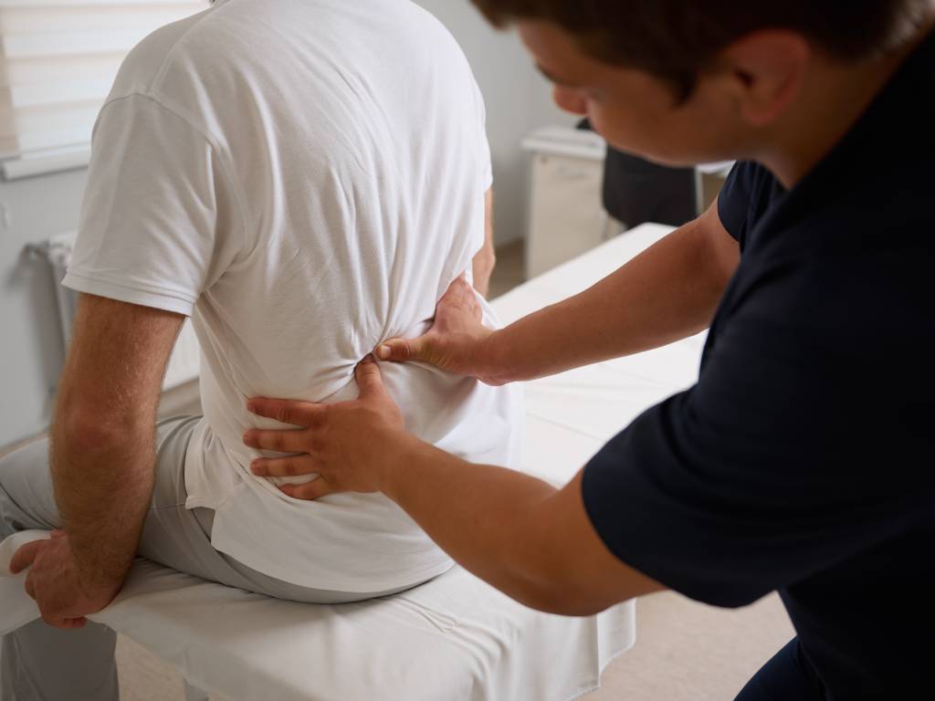 A view from the side and behind of a patient in a white t-shirt sitting on a white medical examination table. A physical therapist or chiropractor wearing dark scrubs is standing next to them, pressing both hands into the patient's lower back to apply targeted pressure to the lumbar region.