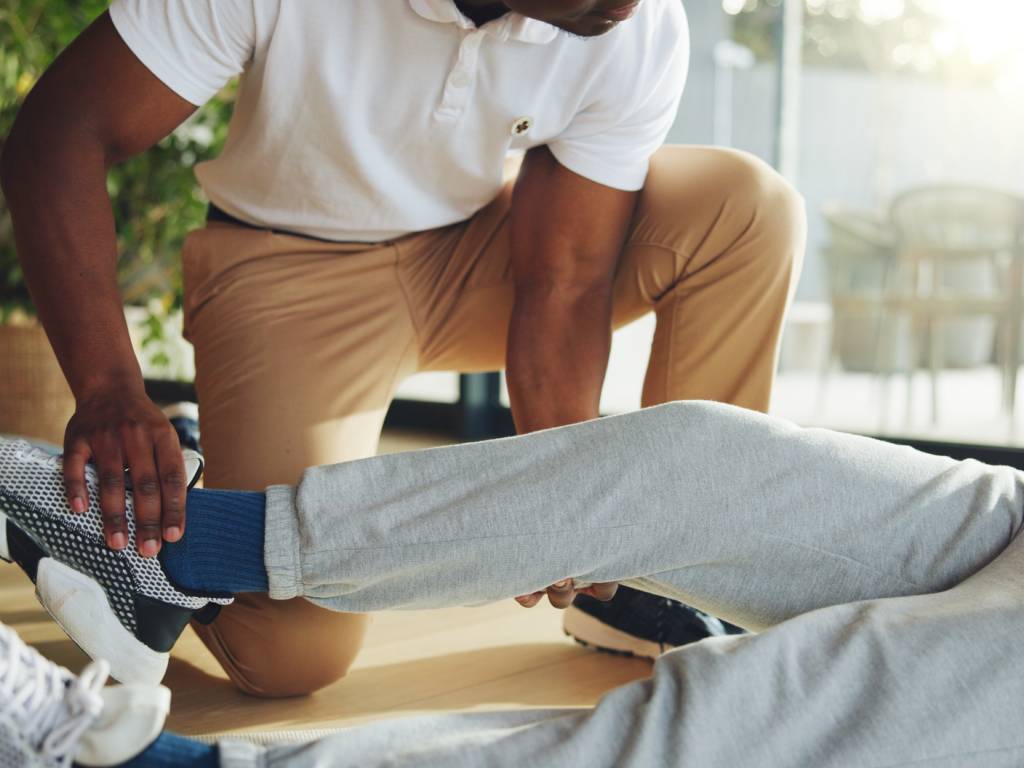 A physical therapist or personal trainer wearing a white polo shirt and khaki pants crouches on a wooden floor to assist a patient lying down. The practitioner holds the patient's ankle and supports under their knee to carefully guide a lower limb stretch, while the patient is dressed in grey sweatpants and patterned sneakers.