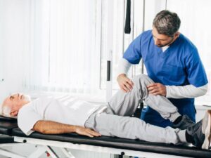 A senior man with white hair lies on a black medical examination table while wearing a white t-shirt and grey sweatpants. A male physiotherapist in blue scrubs stands over him, carefully holding and guiding the patient's bent right knee to assess joint mobility in a bright clinical setting.
