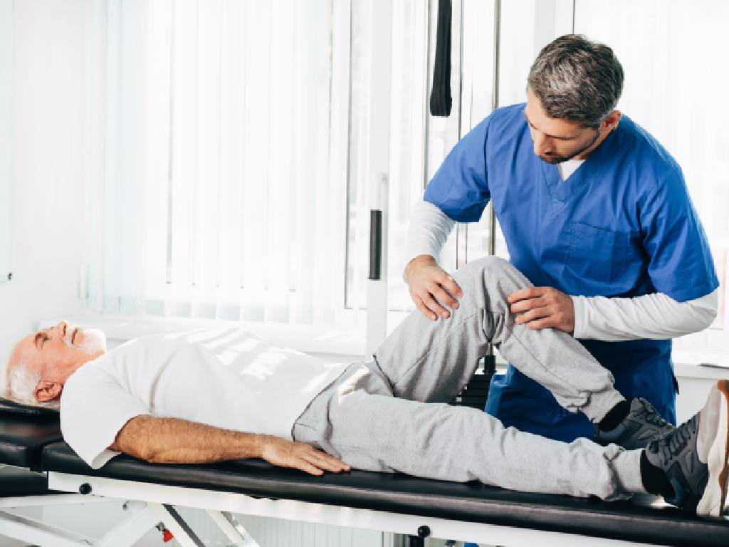 A senior man with white hair lies on a black medical examination table while wearing a white t-shirt and grey sweatpants. A male physiotherapist in blue scrubs stands over him, carefully holding and guiding the patient's bent right knee to assess joint mobility in a bright clinical setting.
