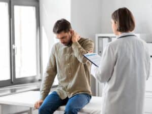 A man wearing a green button-down shirt and blue jeans sits on a white medical examination table, holding the back of his neck with a pained expression. In the foreground, a female healthcare professional in a white lab coat holds a clipboard and listens to the patient in a bright office setting.