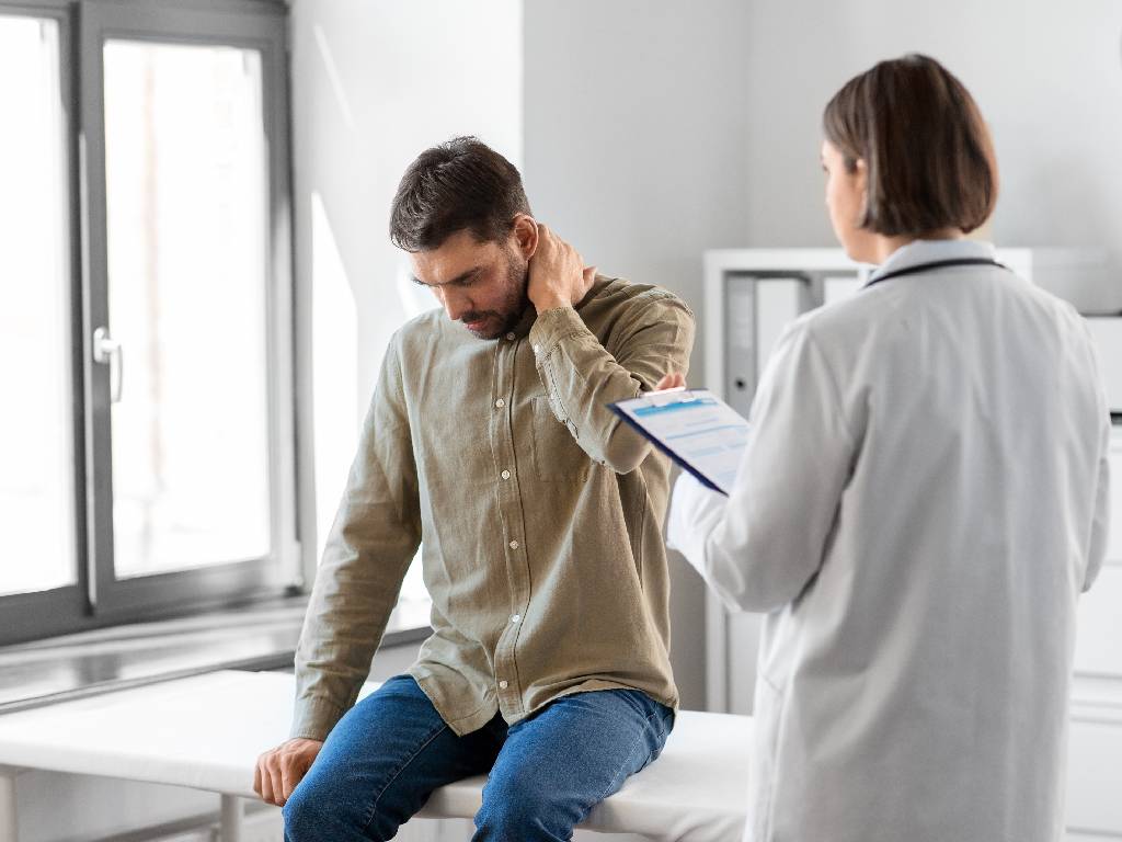 A man wearing a green button-down shirt and blue jeans sits on a white medical examination table, holding the back of his neck with a pained expression. In the foreground, a female healthcare professional in a white lab coat holds a clipboard and listens to the patient in a bright office setting.