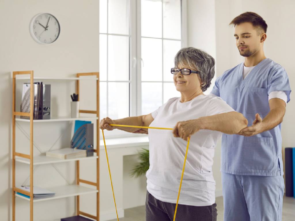 An elderly woman with short grey hair and glasses smiles while performing an upper-body exercise, pulling a yellow resistance band taut across her chest. A male physiotherapist wearing blue scrubs stands closely behind her, gently supporting her elbow and arm to guide her form in a bright clinical setting.