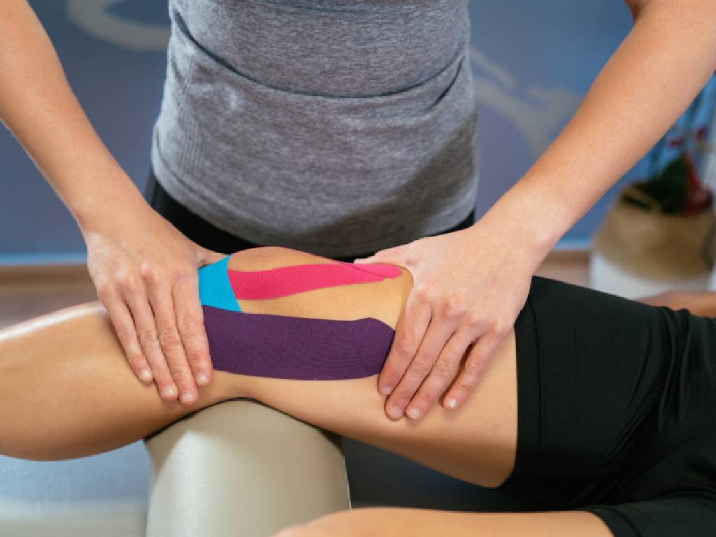A close-up shot of a rehabilitation session where a practitioner in a grey athletic top places both hands firmly around a patient's lower thigh and knee. The patient's leg is elevated on a beige supportive bolster, showcasing colorful strips of kinesiology tape in pink, purple, and blue applied to the muscles surrounding the knee joint.