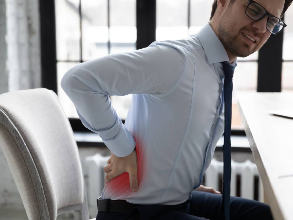 A man in a light blue dress shirt, tie, and glasses winces in discomfort while attempting to stand or sit at his office desk. He has his hand pressed firmly against his lower back and hip, which features a red graphic overlay highlighting the source of his sharp back pain or sciatica.
