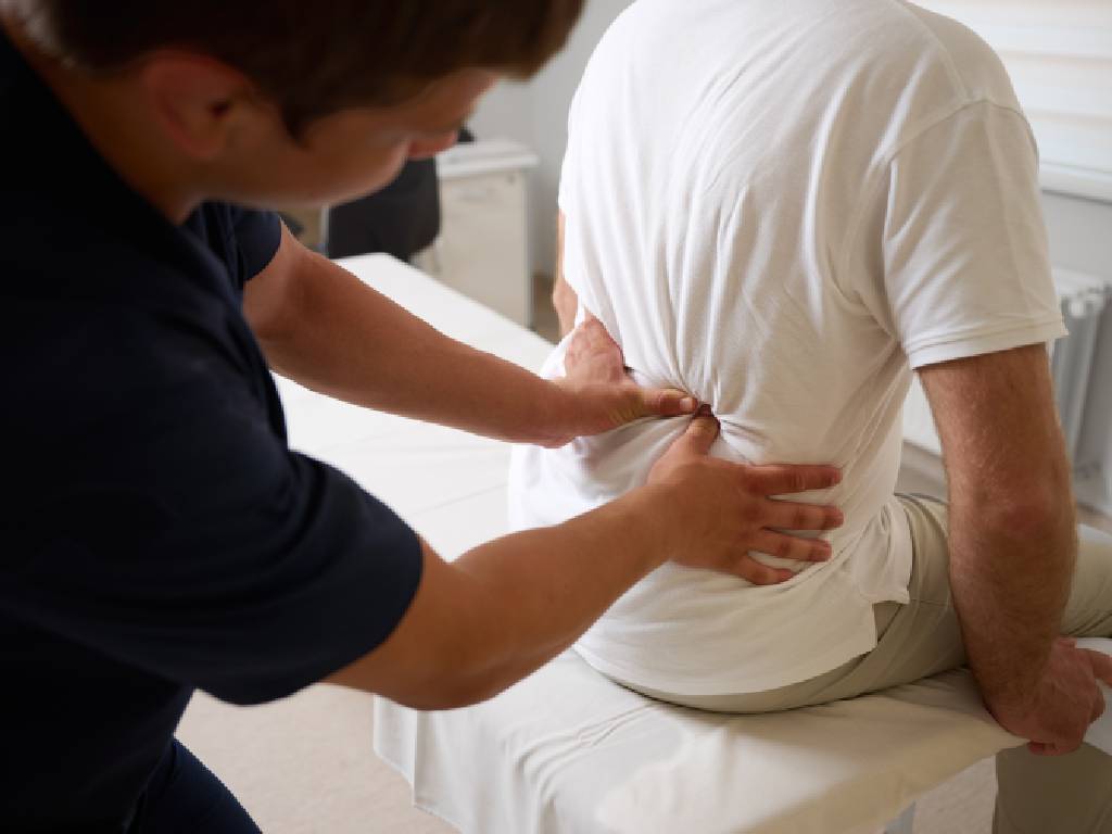 A close-up view from behind a healthcare professional in dark scrubs who is applying targeted manual pressure to the lumbar region of a patient. The patient, wearing a white t-shirt, is seated on a white padded examination table during a chiropractic or physical therapy session.