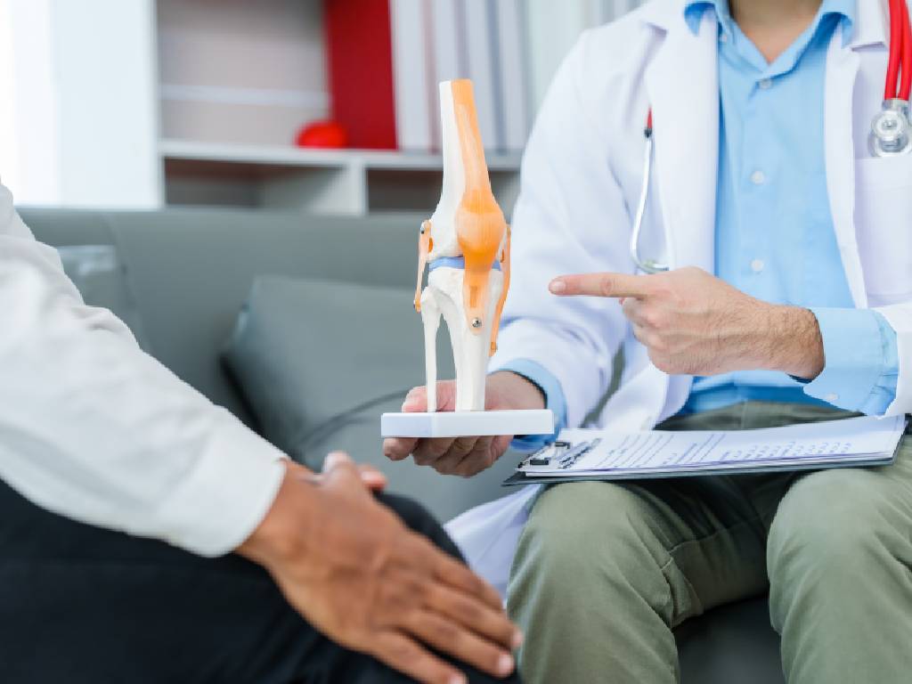 A close-up shot of a medical consultation where a professional in a white lab coat holds a clipboard and points to an anatomical model of a human knee joint, explaining a condition to a patient sitting opposite them.