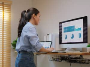 A woman with her hair in a ponytail stands at an adjustable standing desk, looking closely at a large computer monitor displaying financial data charts. She is typing on a keyboard while analyzing graphs, illustrating a modern home office setup and ergonomic workspace posture.