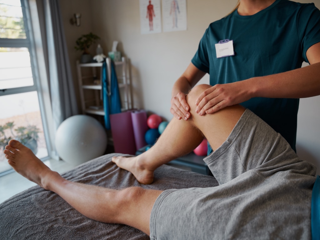 A practitioner wearing a teal scrubs shirt examines a patient's knee on a treatment table in a clinic setting. The therapist's hands are gently placed on the knee joint to assess mobility or joint alignment, illustrating a hands-on physical therapy or chiropractic evaluation.